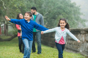 A family of four talking a walk in the park. The two children are walking in front of their parents, smiling and playing with their arms stretched out to the side.