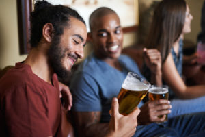Two men sit side by side enjoying glasses of beer. One man smiles at his glass.