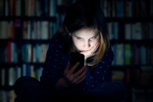 Preteen sits in dark, holding phone, with face lit up by the screen