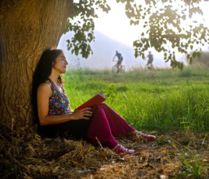 Person with long curly hair sits under tree, relaxed, eyes closed, holding open book on lap