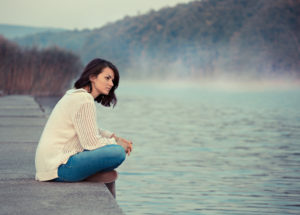 Young adult with shoulder-length hair sitting by lake in the early morning, looking across water