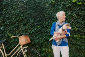 Mature adult with short hair wearing summer scarf holds small dog while standing by bike