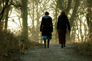 Rear view photo of two people in long skirts and cold-weather wear walking dog along forest path