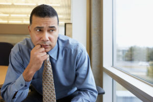 Mature adult in business attire and tie sits on chair in office with worried, thoughtful expression