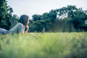 Young person with shoulder-length hair lies on stomach in park using laptop computer