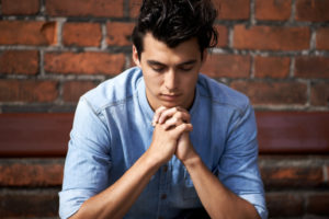 Young adult sits on bench outside brick wall, praying with eyes closed