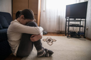 Young teen with ponytail sits on floor alone in living room, head resting on knees