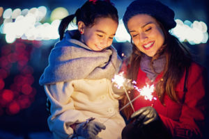 Nighttime scene of adult with long hair and red coat holding sparkler with young child with long hair, hat, and cape