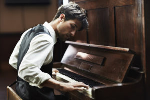 Pianist with short hair wearing vest and dress shirt sits at piano intent on playing