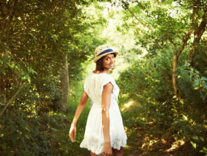 Young adult wearing dress and matching summer hat stands on forest path looking back and smiling