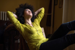 Young person with natural hair wearing green blouse leans back in chair, thoughtfully looking up 