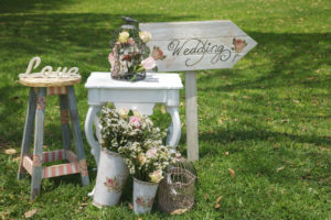 Small table, buckets of flowers, and sign with arrow pointing to the right that reads "Wedding" decorate green lawn