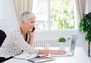 Mature adult with short silver hair sits at desk working on laptop and smiling while talking on phone