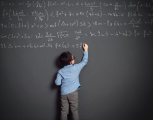 A boy in a blue shirt writes a math equation on a large blackboard.