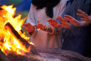 Three people warm their hands over a campfire.