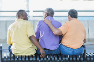 Rear-view of three senior men sitting on a bench. The man on the right has a hand on the middle person's shoulder.