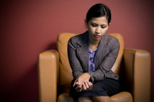 Young adult with hair tied back in ponytail looks down at folded hands, seeming worried