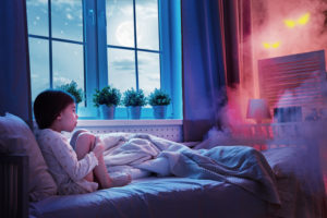 A child sits in bed with knees up to chest, looking afraid of glowing eyes at the end of the bed