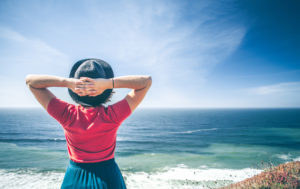 Rear view of person wearing dress and hat looking out to sea with hands behind head