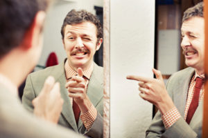 A mustached man winks and points at his reflection in a mirror.