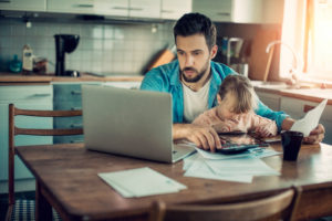 Adult with short hair wearing casual clothes sits at table and works from home with toddler on lap