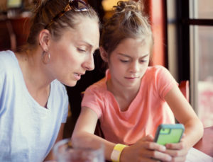 Concerned parent with hair tied back leans over young preteen's shoulder and looks at what teen is doing on smartphone