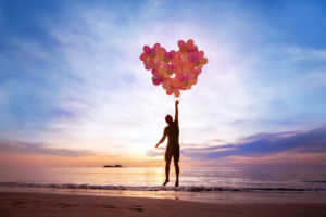 Person at beach holds heart-shaped bunch of balloons and rises slightly off ground