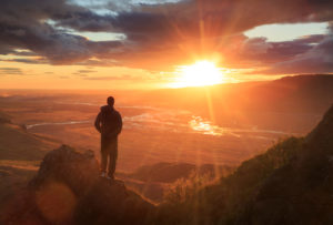 Person stands on a ledge of a mountain, enjoying the sunset over a river valley 