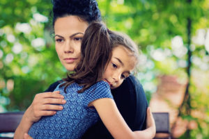 Parent with determined expression holds child on lap on park bench outside. Child leans toward her but looks away