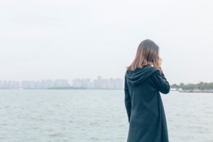 Person with shoulder-length hair wearing long dark sweater stands alone at riverside having phone conversation. 