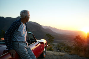 A senior man leans against his car as the sun rises.