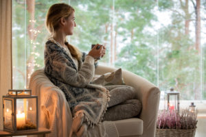 Person with long hair in braid sits looking out window holding hot drink wrapped in sweater