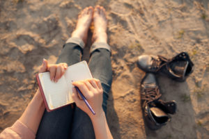 Photo of legs and feet of person who is holding a journal on lap and writing on sandy beach