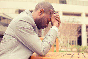 Person dressed in business suit sits outside at table and rubs forehed