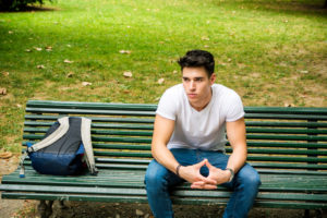 Young student with short hair sits on bench, looking lonely, backpack nearby
