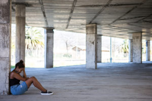 Teenager with hair in ponytail sits in stone building looking down alone