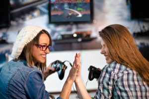 Two women hi-five each other. They each hold a video game controller in their free hand.