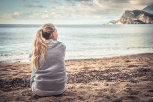 Thoughtful young adult with long hair in ponytail sits on beach and looks out over water