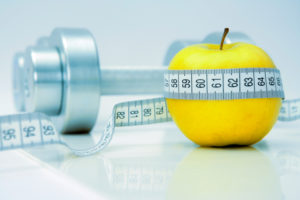 Close-up of an apple with a measuring tape around it and chrome dumbbells at the background. 