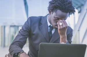 A man in a suit pinches the bridge of his nose. He is using his laptop outdoors.