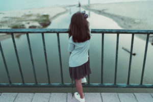 Child wearing skirt and sneakers stands at bridge and looks out over water