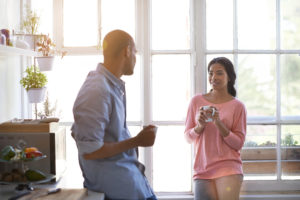 Two adults stand in the kitchen at home, holding mugs and talking with each other