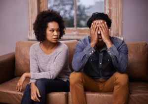 A man and a woman sit together on a brown couch. They both look upset.
