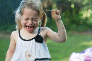 Young child with blonde pigtails wearing white dress cries angrily outside in park