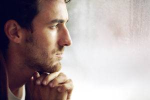 Young adult with short hair and facial hair, hands clasped under chin, looks into distance thoughtfully