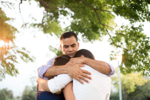 Hispanic parent hugs two teenage children outside under trees 