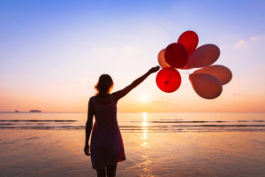 Person looks out to sea, raising arm with bunch of balloons to sky