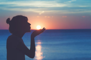 Person with hair tied back holds hand to look as if she is holding the sun that is setting out at sea