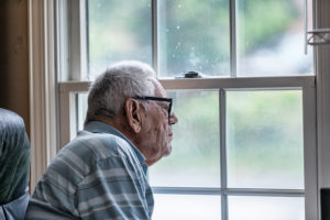 A white-haired man in glasses looks out a dirty window.