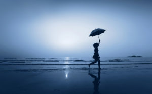 Young adult runs along shore with umbrella against blue-filtered sky